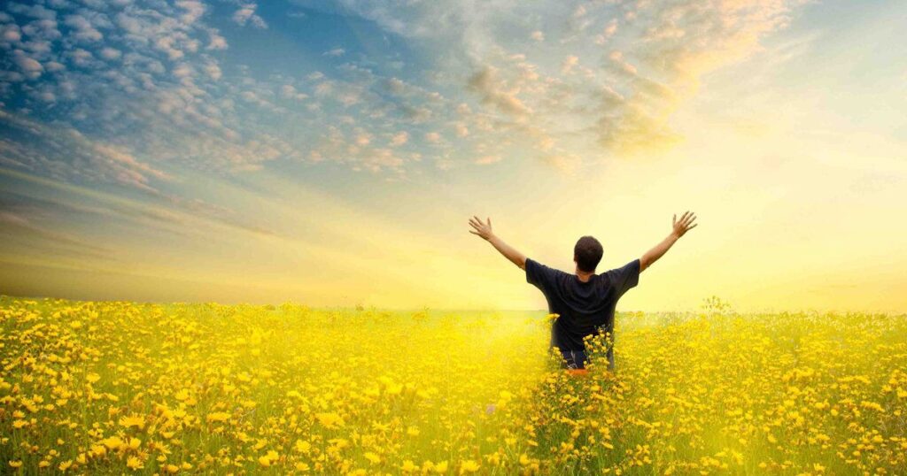 A man stands in a field of yellow flowers, arms outstretched, embodying a moment of reflection in a Short Sunday Morning Prayer.